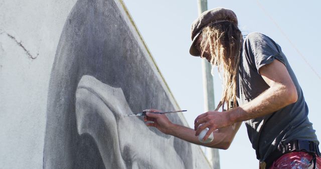 A muralist with dreadlocks painting a monochrome mural on an outdoor wall. The image captures the dedication and skill involved in creating large-scale street art. Suitable for use in articles about street art, creative endeavors, public art projects, and urban culture.
