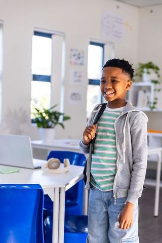 Smiling african american schoolboy with backpack and laptop in bright classroom prepared