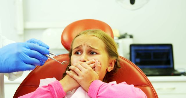 Young girl covering her mouth with hands while dentist in background holding dental tools. Focus is on child's fearful expression. Strong imagery for topics on pediatric dentistry, childhood anxieties, and promoting dental health awareness. Can be used in articles, advertisements for pediatric dental clinics, or informational materials on addressing children's fear of dental visits.