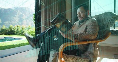 Asian man reading in armchair by glass doors with financial data overlay and pool view