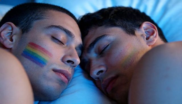 Sleeping male couple cuddling with rainbow face paint on cheeks in blue-lit bedroom