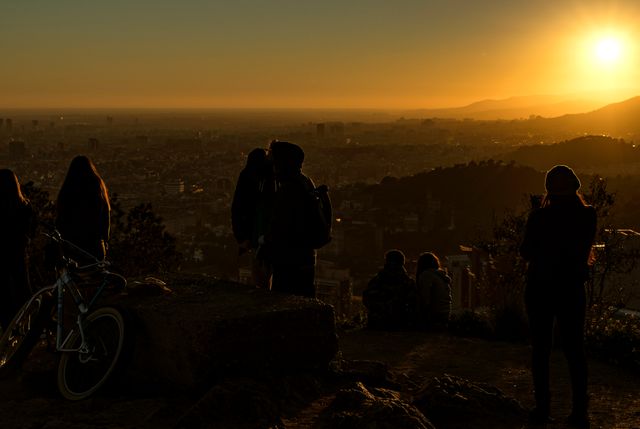 Hikers are enjoying the view of a city at sunset from a hilltop, captured in silhouette against the warm glow of the setting sun. Ideal for travel and adventure concepts, urban and nature contrasts, or outdoor lifestyle visuals.