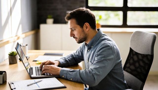 Focused remote worker typing on laptop at tidy home office desk with clipboard