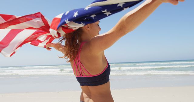 A young woman is holding an American flag while standing on a sunny beach. She appears joyful and energetic, embodying a sense of freedom and patriotism. The ocean waves and clear blue sky enhance the vibrant and uplifting mood. Ideal for use in advertising campaigns for holidays like Independence Day, travel and tourism promotions, and any content celebrating American patriotism or summer beach activities.