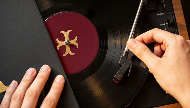Hands lowering tonearm onto vinyl record with maroon label and gold emblem, turntable close-up