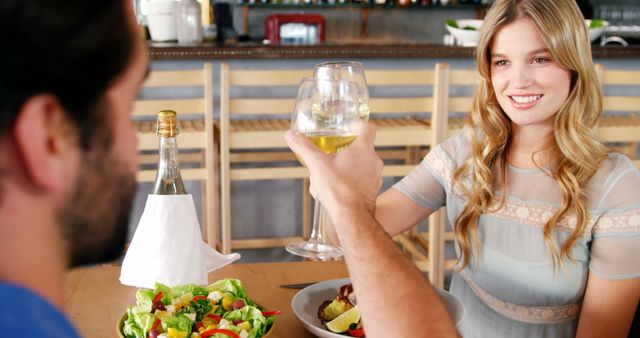 A young Caucasian woman and man are toasting with wine glasses at a dining table, with copy space. Their cheerful engagement suggests a romantic dinner or celebration in a cozy restaurant setting.