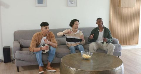 Diverse young men sitting on gray sofa holding bottled drinks enjoying casual game night