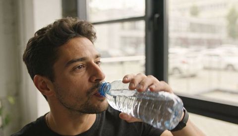 Young man drinking bottled water while wearing smartwatch near window in modern interior