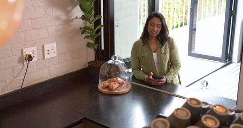 Indian woman wearing olive green jacket checking smartphone at cafe counter with pastries