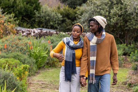 African american couple walking hand in hand in garden wearing mustard and rust sweaters