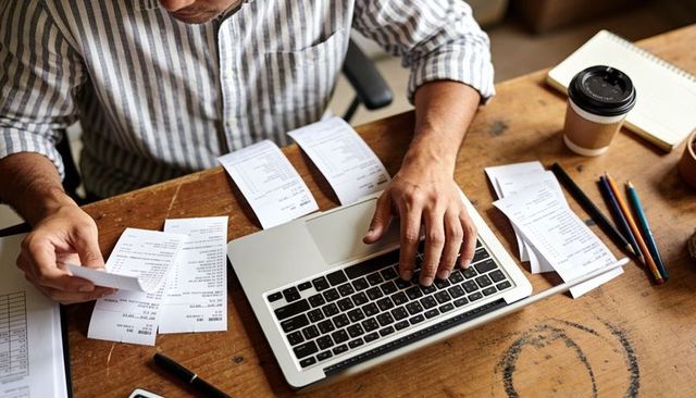 Man sorting receipts entering expense data on laptop at home desk with coffee cup