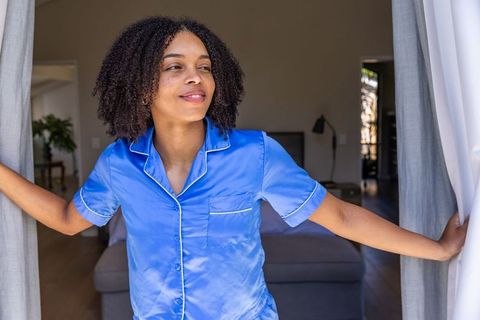 African american woman opening curtains in blue satin pajamas morning routine