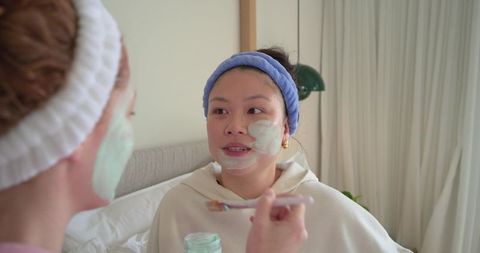 Two diverse women applying mint green face mask on bed during cozy morning skincare ritual