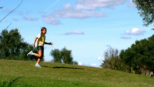 A young girl is running enthusiastically across a grassy park on a sunny day, illustrating the joy and freedom of playing outdoors. The lush green landscape contrasted with clear blue sky creates a serene outdoor vibe. This can be used for promoting healthy outdoor activities for children, nature relaxation themes, or emphasizing a playful experience in a natural setting.