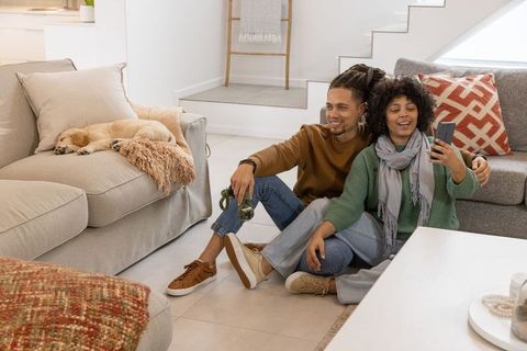 African american couple relaxing on living room floor taking selfie with sleeping puppy