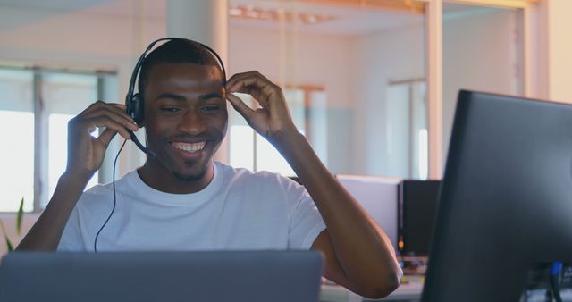 A friendly customer support representative is smiling while wearing a headset and working on a computer in a modern office. This image is perfect for illustrating concepts of professional communication, tech support, customer service, modern workplaces, and work-related articles or promotional materials for business solutions.