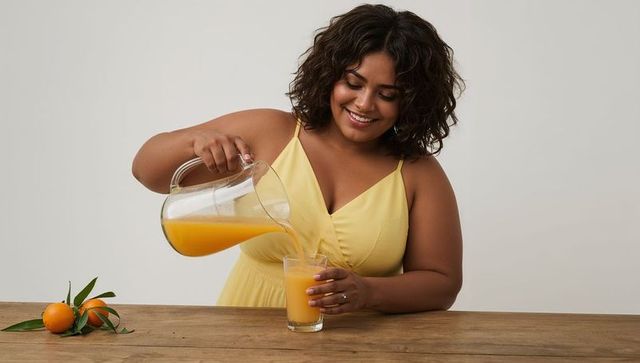 Smiling woman pouring fresh orange juice from glass pitcher into tumbler on wood table