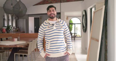 Smiling indian man standing in bright modern open-plan home wearing striped pullover