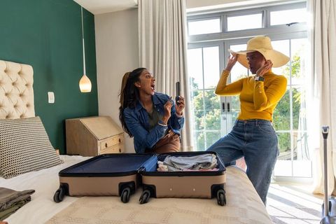 Diverse friends packing luggage and trying straw hat in sunlit bedroom