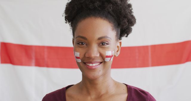 This vibrant photo shows a young African woman smiling confidently with the England flag painted on her cheeks. Ideal for articles and ads related to sports events, national celebrations, team spirit, multiculturalism, and patriotism.
