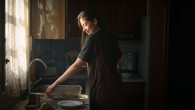 Smiling woman washing dishes in sunlit vintage kitchen wearing plaid pinafore apron