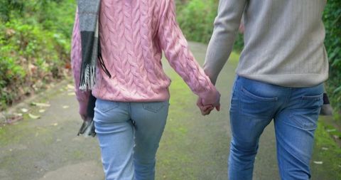 Couple walking hand in hand on mossy park path wearing knit sweaters, holding gloves