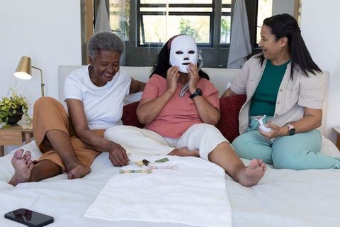 Three women laughing using led facial mask and handheld skincare device on bed