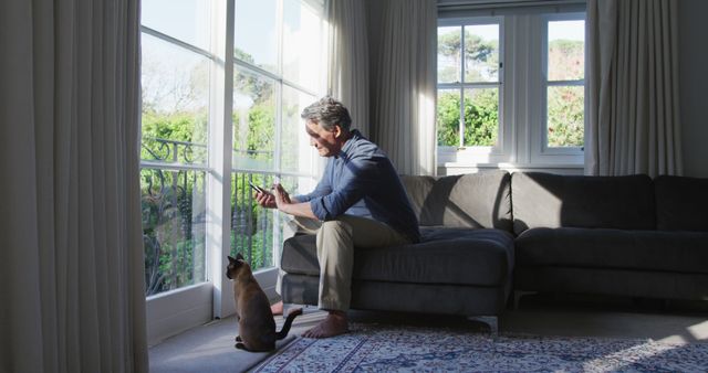 Happy senior caucasian man sitting in sunny living room using smartphone and stroking cat. retirement lifestyle at home.