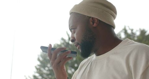 African man speaking into smartphone by window wearing beige beanie and white shirt