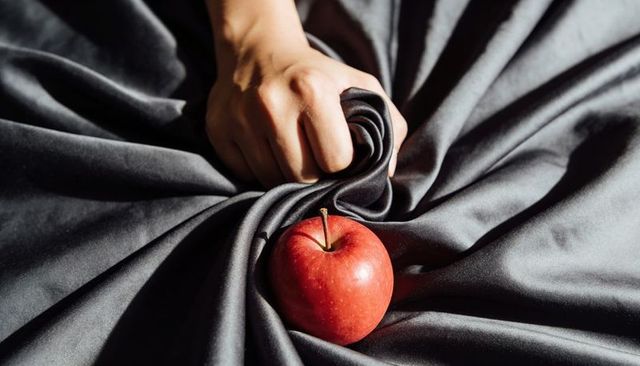 Hand gripping dark satin fabric with glossy red apple for moody texture contrast