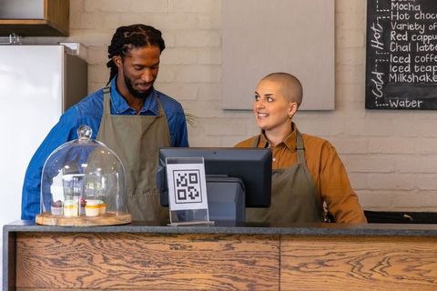 Diverse baristas smiling and using touchscreen pos at cozy wooden coffee counter