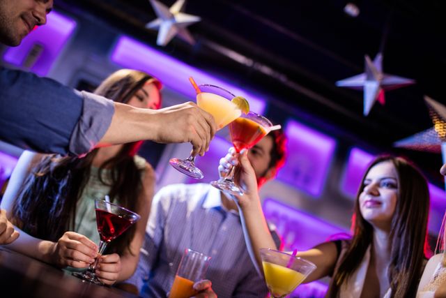 Group of friends raising cocktails in a toast at a trendy bar. Ideal for use in content related to nightlife, social gatherings, celebrations, and young adult lifestyle. Perfect for advertising bars, clubs, and social events.