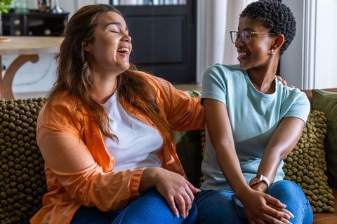 Two african american women laughing and chatting on cozy green couch in sunlit living room