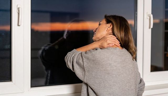 Woman leaning at window, touching neck and gazing at sunset horizon, evening contemplation