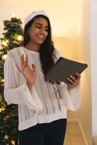 Cheerful Woman with Tablet by Christmas Tree in Festive Santa Hat