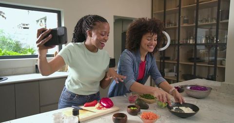Diverse women filming cooking vlog while preparing colorful bowl on marble kitchen island