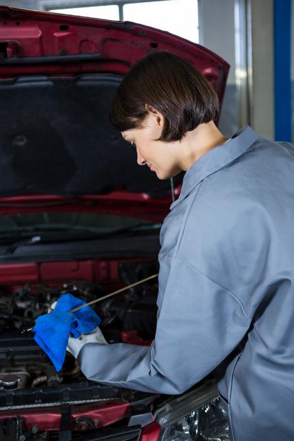Female mechanic checking oil level in car engine using a dipstick at a repair garage. Ideal for illustrating automotive services, gender diversity in technical fields, and professional car maintenance. Useful for websites, brochures, and advertisements related to auto repair shops, mechanical training programs, and vehicle maintenance services.