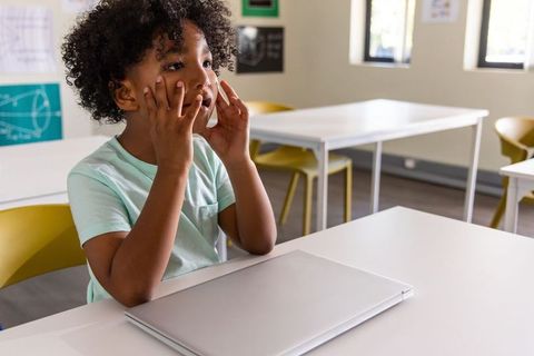 Curious african american child sitting in classroom touching cheeks beside closed laptop
