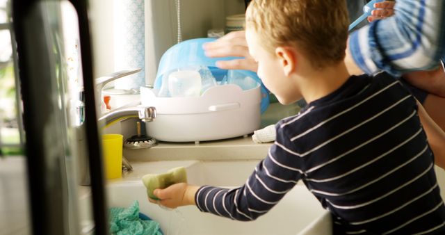 Young boy wearing striped shirt washing dishes in bright kitchen. He seems focused on the task at hand. Green sponge and blue dishtowel are visible. Adult’s hand nearby aiding with the task. Ideal for use in articles about household chores, family teamwork, or parenting tips.