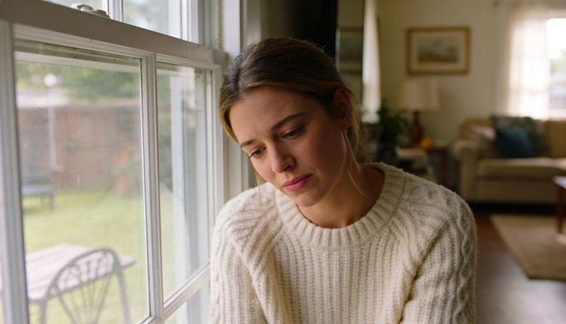 Young woman gazing out window in cream cable-knit sweater, contemplative cozy home portrait