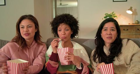 Multicultural women watching movie and eating popcorn on cozy living room couch together