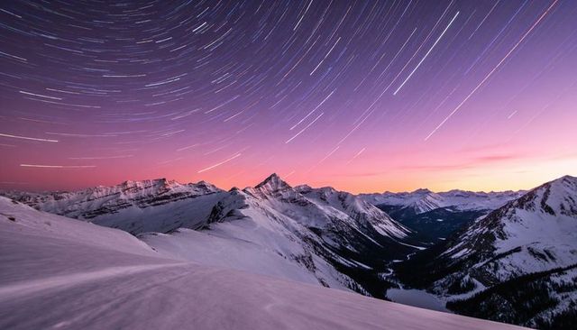Star trails sweeping over snow-capped mountain ridge and frozen valley at twilight