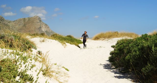 Man running along sandy trail through scenic beach dunes, with mountains and a clear blue sky in the background. Ideal for use in fitness, outdoor, health and travel activities advertisements. Great for illustrating active lifestyles, promoting tourism, and showcasing natural beauty.