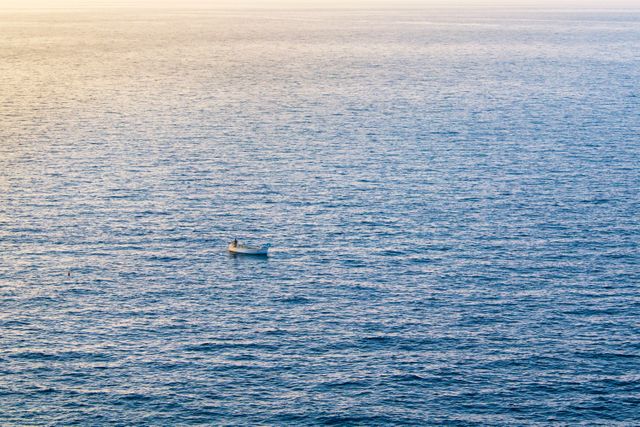 Peaceful scene of a single boat floating on a calm ocean during sunset. The tranquil setting with blue water extending to the horizon makes it an ideal image for themes of solitude, relaxation, and meditation. Perfect for use in travel brochures, websites about oceanic activities, or backgrounds for inspirational quotes.
