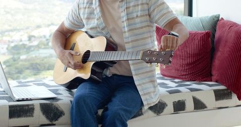 Tuning acoustic guitar on window seat with laptop and city view