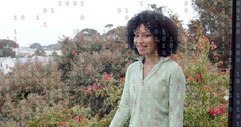 Smiling woman wearing green top standing on balcony with flowering shrubs and binary overlay