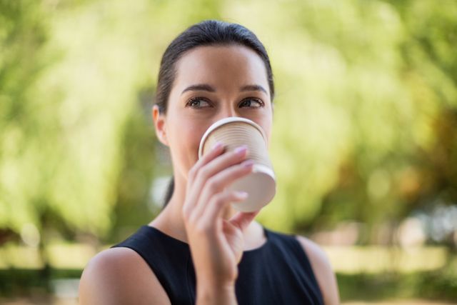 Woman drinking coffee from a disposable cup in an outdoor setting with greenery in the background. Ideal for use in lifestyle blogs, coffee shop promotions, advertisements for outdoor activities, and wellness articles.