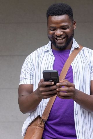 Smiling african american man checking smartphone wearing purple tee and brown satchel