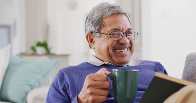 Image of happy senior biracial man drinking coffee, reading book at home and laughing. Retirement, hobbies, domestic life, inclusivity and senior lifestyle concept.