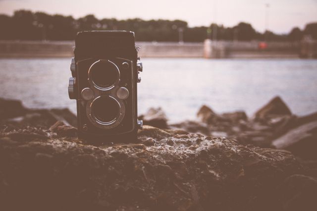 This image features a vintage twin-lens reflex camera placed on rocks near a body of water, possibly a river or lake. The warm tones and soft focus give it a nostalgic and retro feel. It is ideal for use in content related to classic photography, retro-themed designs, or hobbyist photography topics.